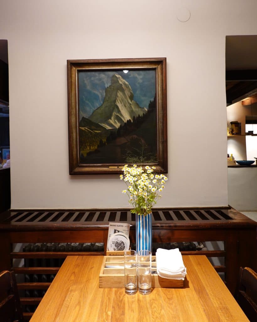 Een houten tafel met een blauw gestreepte vaas met madeliefjes, glazen, servetten en een menukaart staat onder een ingelijst Tirools bergschilderij op een witte muur in een gezellige, warm verlichte kamer.