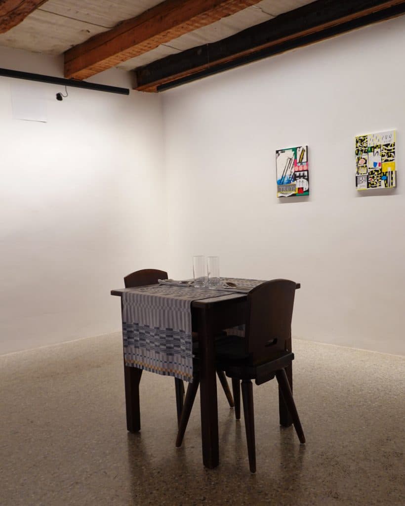 Een kleine houten tafel met twee stoelen is gedekt met lege glazen kopjes en een geruite tafelloper in een minimalistische kamer met witte muren en zichtbare balken, perfect voor je Tirol zomerchecklist. Twee kleurrijke abstracte kunstwerken hangen erachter.