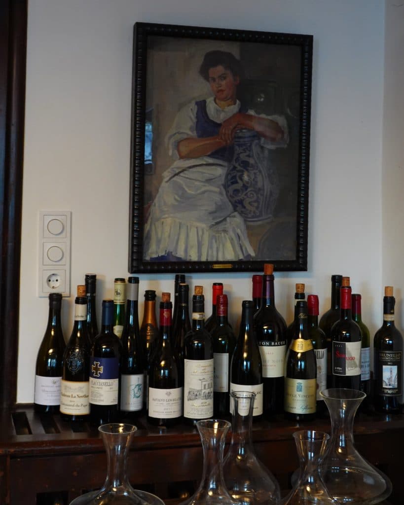 Een groep verschillende wijnflessen staat op een houten plank onder een ingelijst portret van een zittende vrouw in een witte jurk, perfect voor je zomerchecklist. Drie glazen karaffen staan voor de flessen.