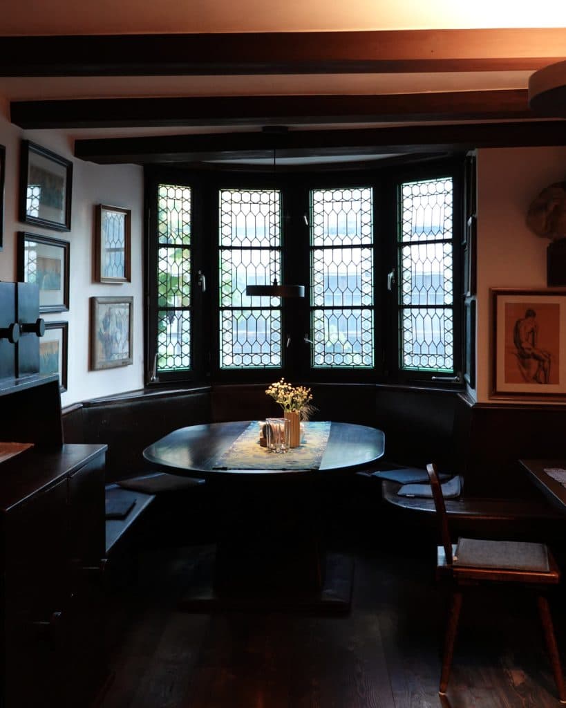 Een gezellige eethoek met een ronde houten tafel, een bankje en een vaas met bloemen staat bij grote tralievensters. Natuurlijk licht valt binnen en ingelijste kunstwerken geïnspireerd door Tirol sieren de witte muren. Perfect voor je Zomerchecklist.