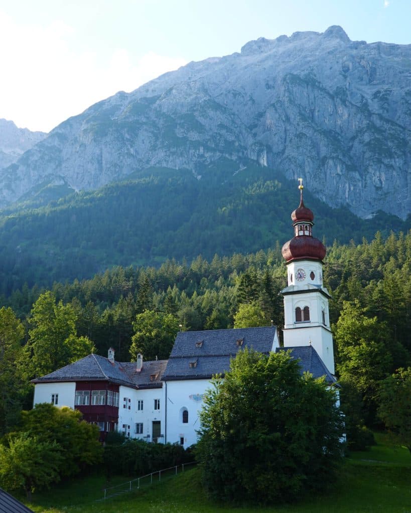Een witte kerk met een rode uivormige koepel staat tussen de bomen in Tirol, met steile beboste bergen die dramatisch oprijzen op de achtergrond.