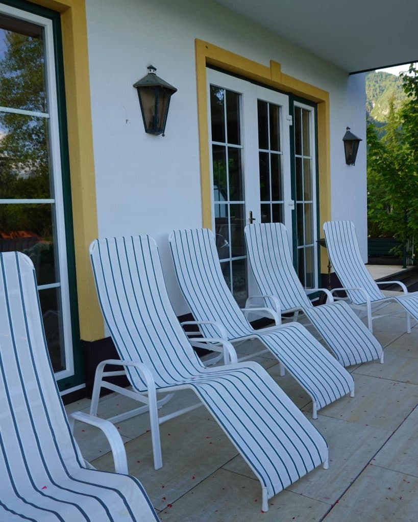 Vijf witte loungestoelen met blauwe strepen staan in een rij op een betegelde patio voor glazen deuren en ramen van een wit gebouw met gele sierstrips. Dit is de perfecte plek om te ontspannen na het invullen van je Zomerchecklist voor een uitje in Tirol.