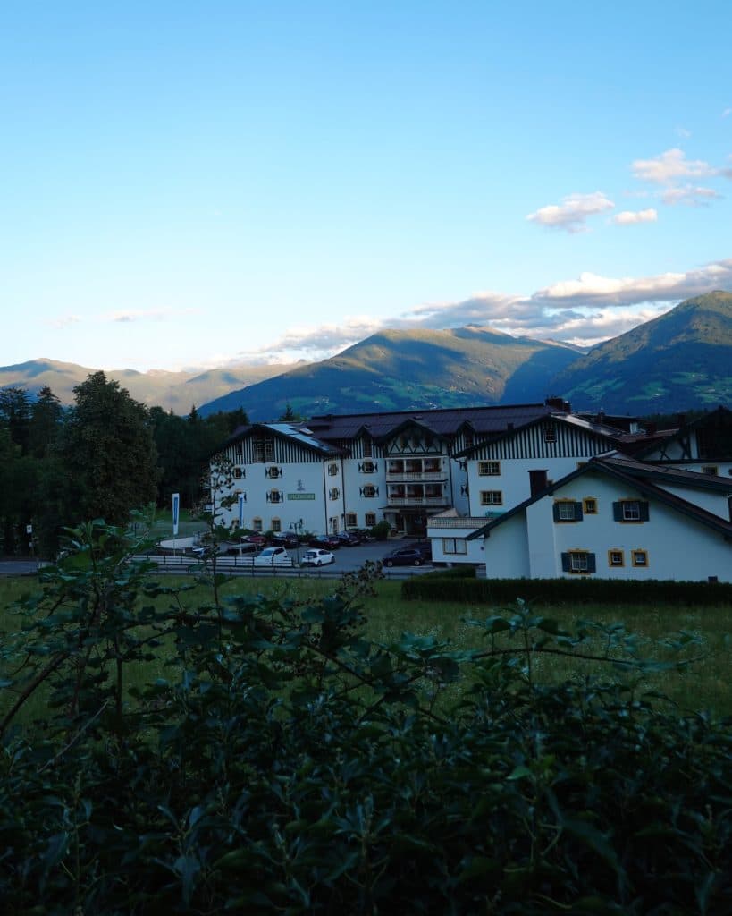 Een wit hotel in alpinestijl met donkere daken ligt aan de voet van de groene bergen van Tirol onder een blauwe lucht met verspreide wolken. Struiken en gras staan op de voorgrond en het zonlicht verlicht de bergen in de verte.