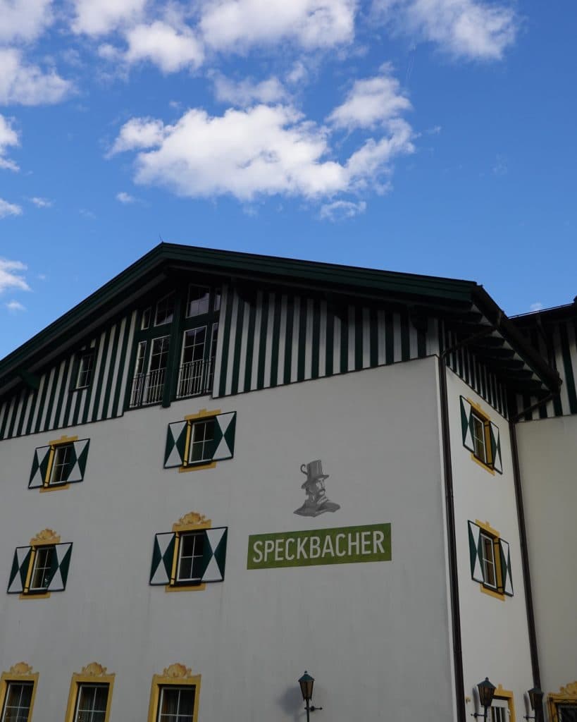 Een wit gebouw in Tirol met groene en gele luiken en een bord met de tekst "SPECKBACHER" onder een afbeelding van een hoed, tegen een blauwe lucht met verspreide wolken - perfect voor je zomerchecklist.