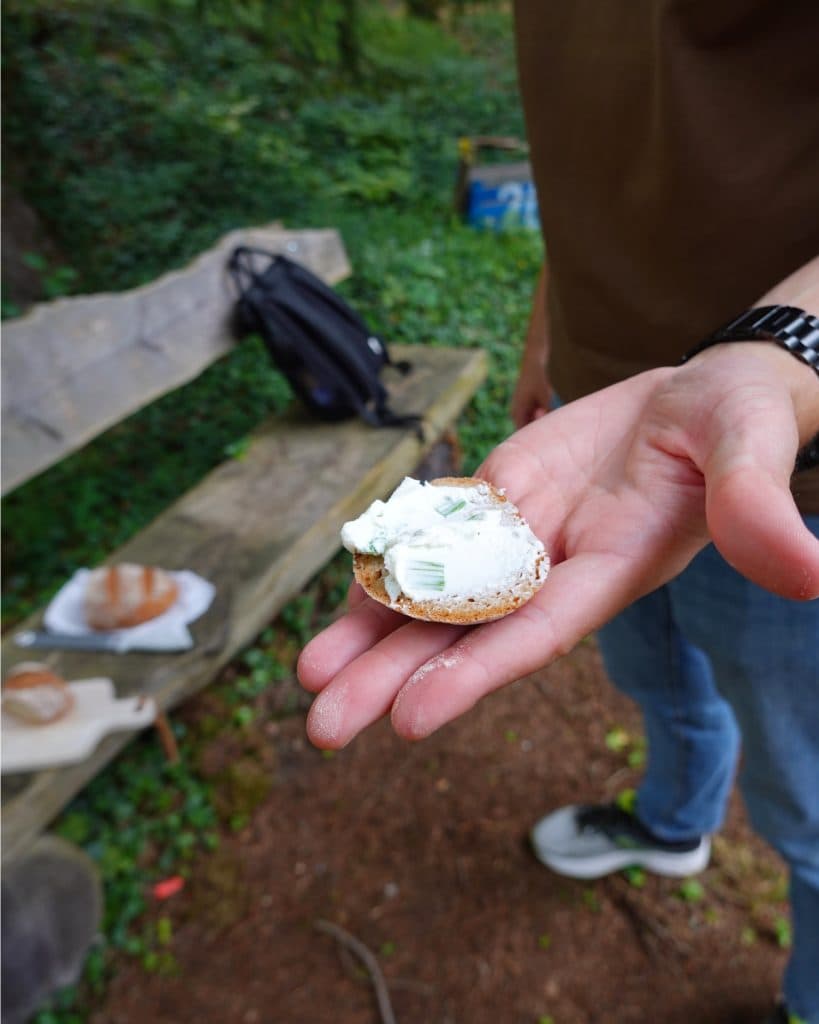 Een persoon met een ronde cracker belegd met roomkaas en kruiden geniet van de buitenlucht in Tirol. Op de achtergrond staan een houten bankje, een rugzak en een bord met nog meer eten - allemaal op een bladerrijke ondergrond, perfect voor elke Zomerchecklist.
