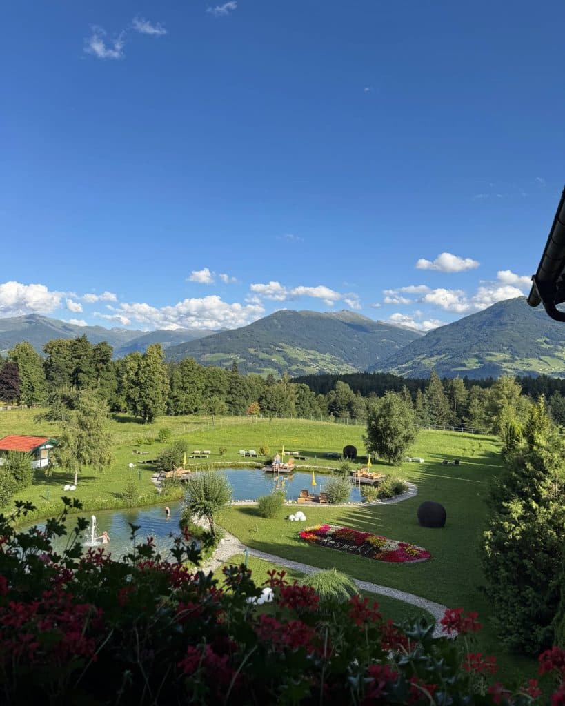 Een weelderig groen landschap met een vijver, rode bloemen op de voorgrond en mensen die ontspannen in een tuin. Op de achtergrond rijzen de heuvels van Tirol en de bergen in de verte op onder een helderblauwe hemel - met uitzichten die perfect zijn voor je zomerchecklist.