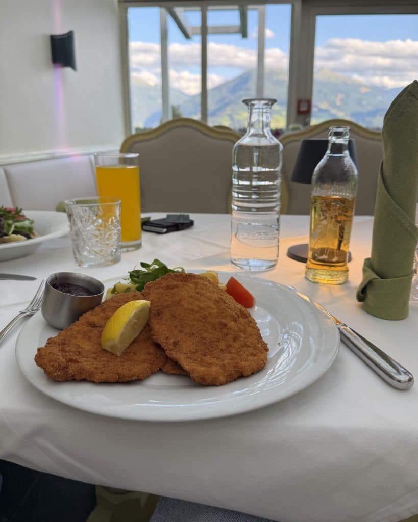 Een bord met twee gepaneerde schnitzels, een citroenpartje, saus en garnering ligt op een wit tafelkleed. Drankjes en bestek zijn netjes gerangschikt terwijl je door de grote ramen geniet van de adembenemende bergen van Tirol - een waar hoogtepunt voor elke Zomerchecklist.