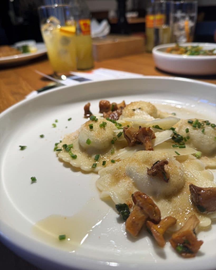 Een close-up van een wit bord met ravioli, belegd met cantharellen en gehakte bieslook, staat op een houten tafel - perfect voor je zomerchecklist na een wandeling in Tirol. Op de achtergrond zijn vage drankjes en een ander gerecht zichtbaar.
