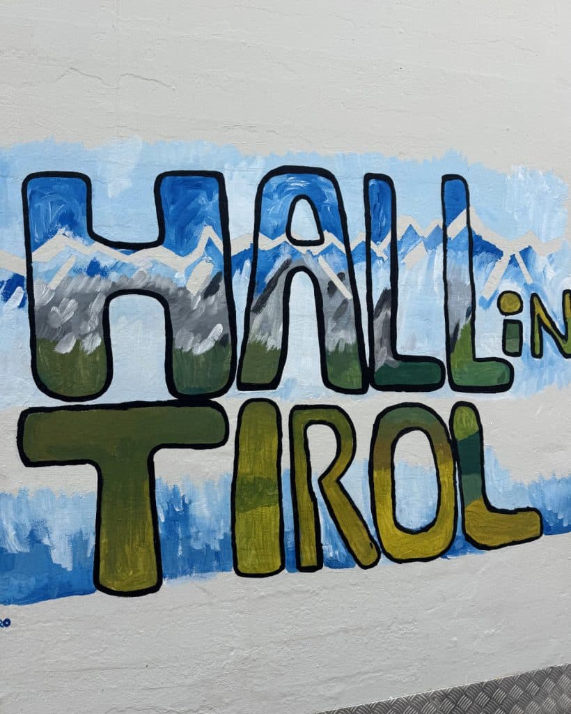 Een muurschildering met grote, kleurrijke letters die "HALL in TIROL" spellen tegen blauwe en witte bergen. Dit handgeschilderde kunstwerk in groen, blauw en geel is perfect voor je zomerchecklist en geeft de levendige sfeer van Tirol weer.