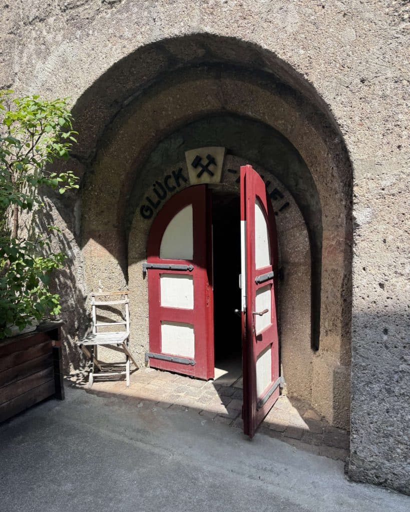 Een stenen boog met open rode en witte dubbele deuren leidt naar een schemerige tunnel in Tirol. Boven de ingang zie je het woord "GLUCK" en twee gekruiste hamers. Buiten staat een houten stoel - perfect om je zomerchecklist door te nemen.