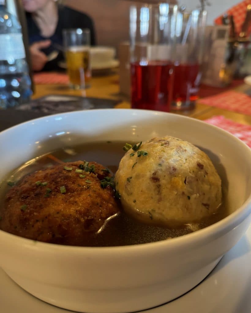 Een witte kom heldere soep met twee grote knoedels, een gebruinde en een lichte, gegarneerd met kruiden - precies zo'n troostrijk gerecht als je in Tirol zou aantreffen. Op de achtergrond glazen rode drank, een biertje en een persoon die gedeeltelijk zichtbaar is.