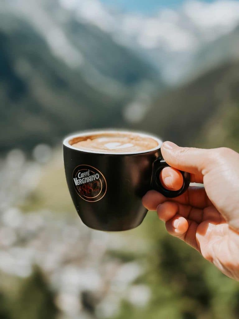 Een hand die een zwarte Caffè Vergnano-kop gevuld met koffie vasthoudt, met op de achtergrond de Bergenmagie van het vage berglandschap van Valle d'Aosta.