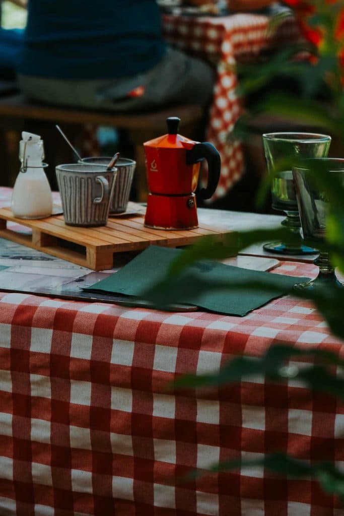 Op een tafel met een rood en wit geruit tafelkleed staan een rode mokapot, een grijze mok met lepel, een kleine witte kan en groene glazen. Dit geeft de gezellige charme weer van een roadtrip in Valle d'Aosta. Op de achtergrond staan vage planten en een andere tafel.