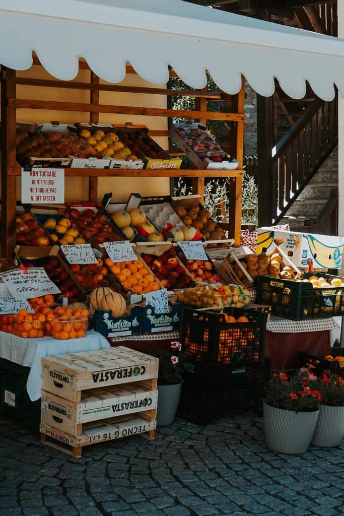 Een kleurrijke fruitkraam in de openlucht toont verschillende soorten fruit in kratten en manden onder een witte luifel, met borden in het Italiaans en bloemen in potten ervoor op een geplaveide straat - perfect voor een roadtrip in Valle d'Aosta.