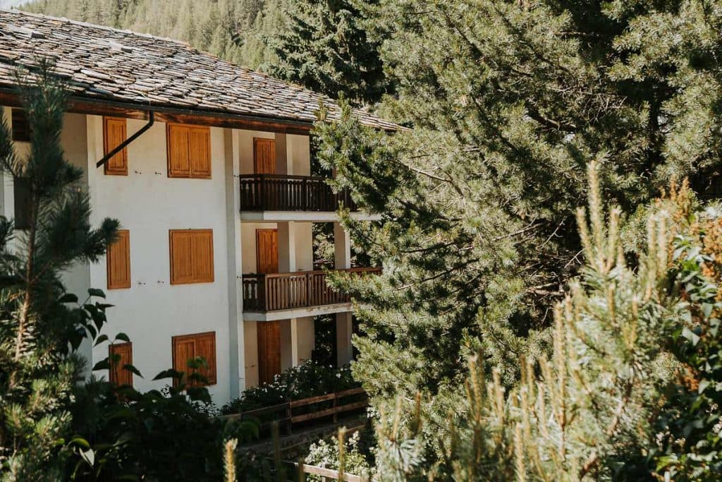 Een wit gebouw met gesloten houten luiken en balkons wordt gedeeltelijk verborgen door hoge, groene pijnbomen in een bosrijk gebied op een zonnige dag-een vredige stop tijdens een roadtrip in Valle d'Aosta.