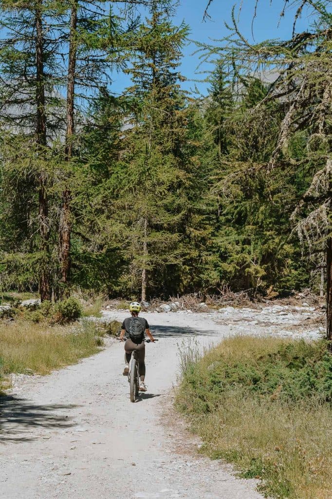 Een persoon met een helm op fietst op een onverhard pad door een bos met hoge groene bomen onder een strakblauwe hemel, genietend van een schilderachtige roadtrip in Valle d'Aosta.