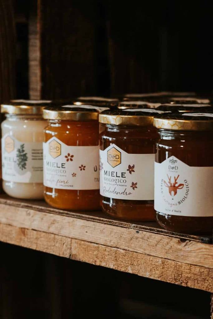 Potten honing met gouden deksels en verschillende etiketten staan netjes gerangschikt op een houten plank, wat de charme oproept van een roadtrip in Valle d'Aosta. Op de etiketten staan bloemmotieven en Italiaanse tekst die de verschillende honingsoorten aanduiden.