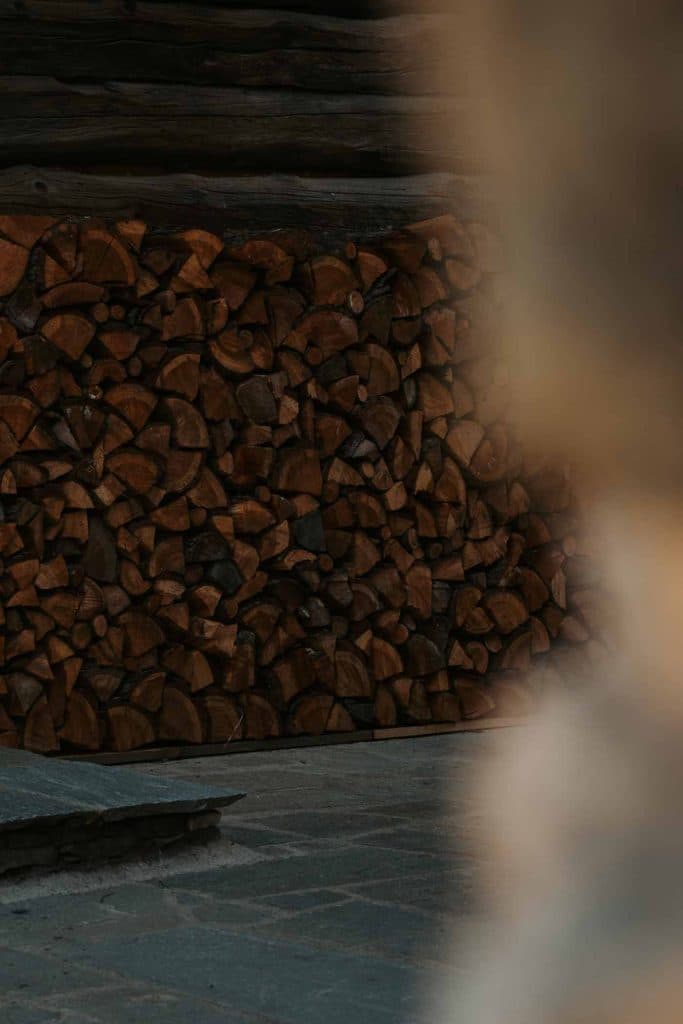 Een netjes opgestapelde stapel brandhout ligt tegen een houten muur, aan de rechterkant gedeeltelijk verborgen door een wazig object op de voorgrond. De schemerige verlichting voegt een warm, rustiek tintje toe - perfect voor een gezellige stop tijdens een roadtrip in Valle d'Aosta.