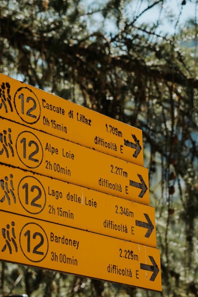 Vier gele wandelroutebordjes in het Italiaans, elk gemarkeerd met het nummer 12 en pijlen die naar rechts wijzen, geven routes, afstanden, looptijden en moeilijkheidsgraden aan - de perfecte leidraad voor een roadtrip door de schilderachtige bossen van Valle d'Aosta.