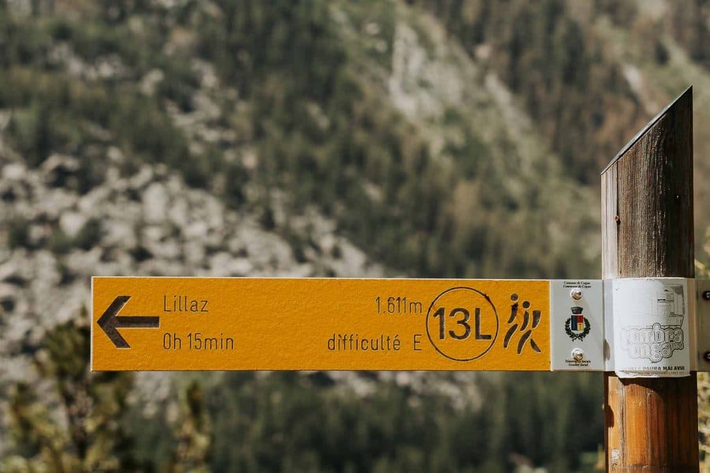 Een geel wandelpadbord wijst naar links in de richting van Lillaz, wat een wandeling van 15 minuten aangeeft, een afstand van 1,61 km en een "E" (gemakkelijk) moeilijkheidsgraad - perfect voor wie op uitstap is in Valle d'Aosta. Padnummer 13L leidt langs vage bomen en rotsachtige heuvels.