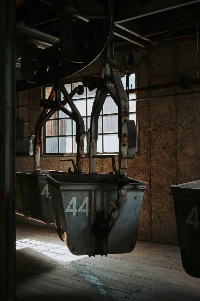 Metalen mijnkarretjes met het nummer 44 hangen aan bovenleidingen in een oud, schemerig verlicht industrieel gebouw - zonlicht dat door een raam naar binnen schijnt wijst op een roadtrip in het rijke industriële verleden van Valle d'Aosta.