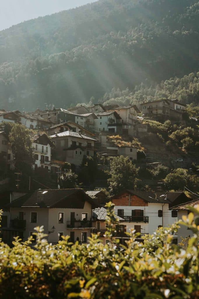 Zonlicht stroomt door bomen naar huizen op een heuvel in een klein dorp omgeven door groen en bergen, met planten op de voorgrond.