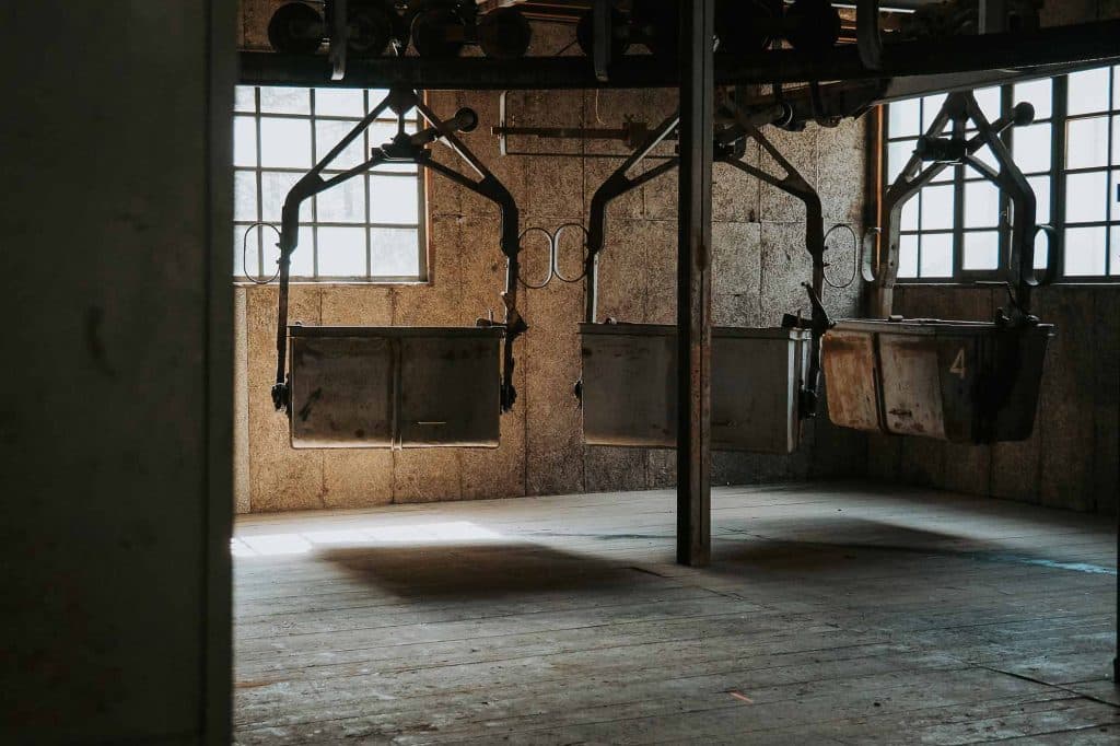 Drie oude, lege mijnkarretjes hangen aan een hangende rail in een schemerig, verlaten industrieel gebouw met houten vloeren en zonlicht dat door twee ramen naar binnen schijnt - een verborgen juweeltje om te ontdekken tijdens een roadtrip in Valle d'Aosta.