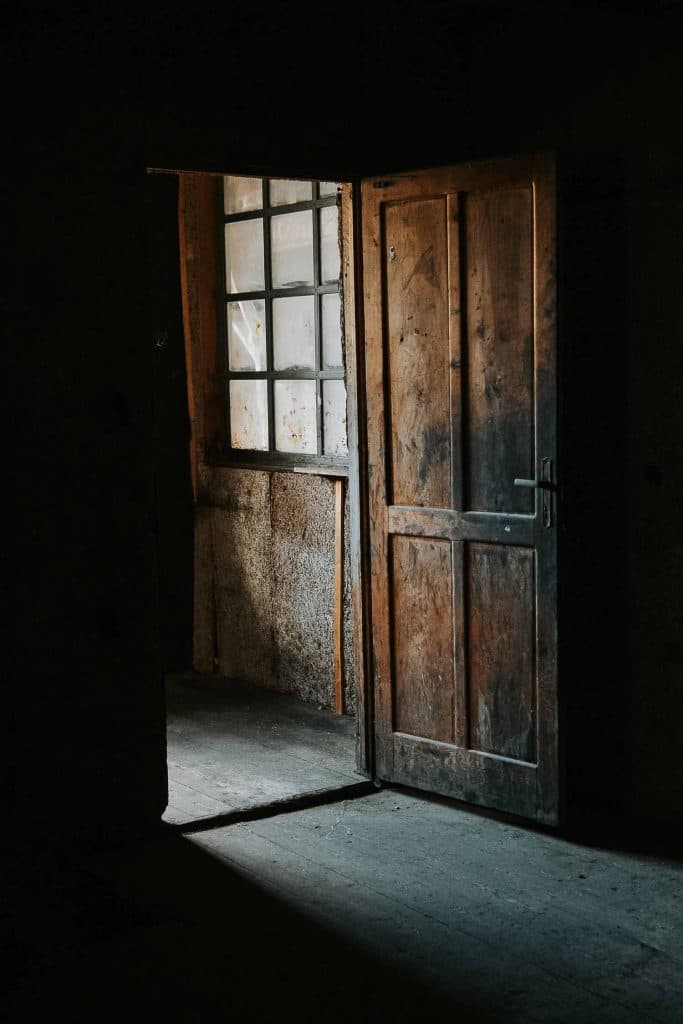 Een houten deur staat op een kier in een zwak verlichte kamer, waardoor er licht naar binnen stroomt dat doet denken aan de vroege ochtenden tijdens een roadtrip in Valle d'Aosta en dat een deel van de ruwe, getextureerde muren en vloer verlicht.