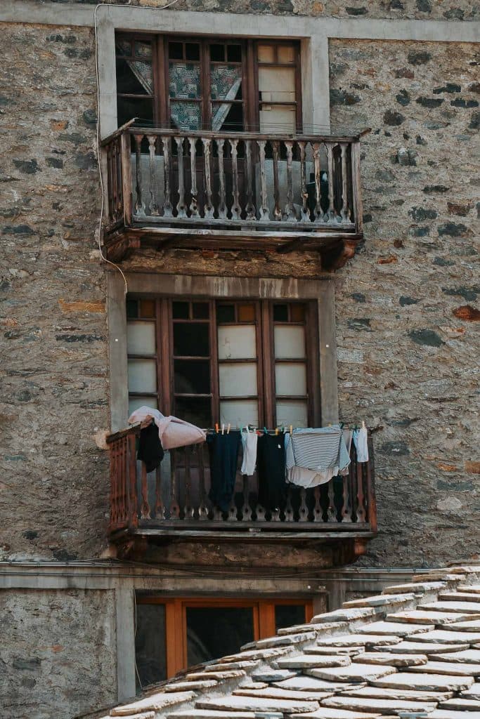 Twee kleine houten balkons op een stenen gebouw; op het onderste balkon hangt wasgoed, waaronder overhemden en broeken, aan een waslijn. Ontdek authentieke details zoals deze tijdens je roadtrip in Valle d'Aosta, waar ramen zijn voorzien van meerdere glazen ruiten.