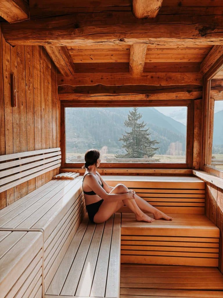 Een vrouw in een zwart badpak zit op een houten bank in een sauna en kijkt uit een groot raam naar bergen en bomen in de verte - een serene stop tijdens haar roadtrip in Valle d'Aosta. Warm natuurlijk licht vult het houten interieur.