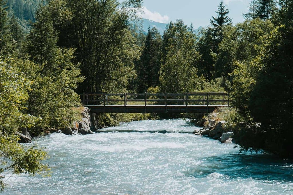 Een houten loopbrug overbrugt een stromende rivier omringd door weelderige groene bomen, met bergen zichtbaar op de achtergrond onder een heldere, zonnige hemel - perfect decor voor een roadtrip in Valle d'Aosta.