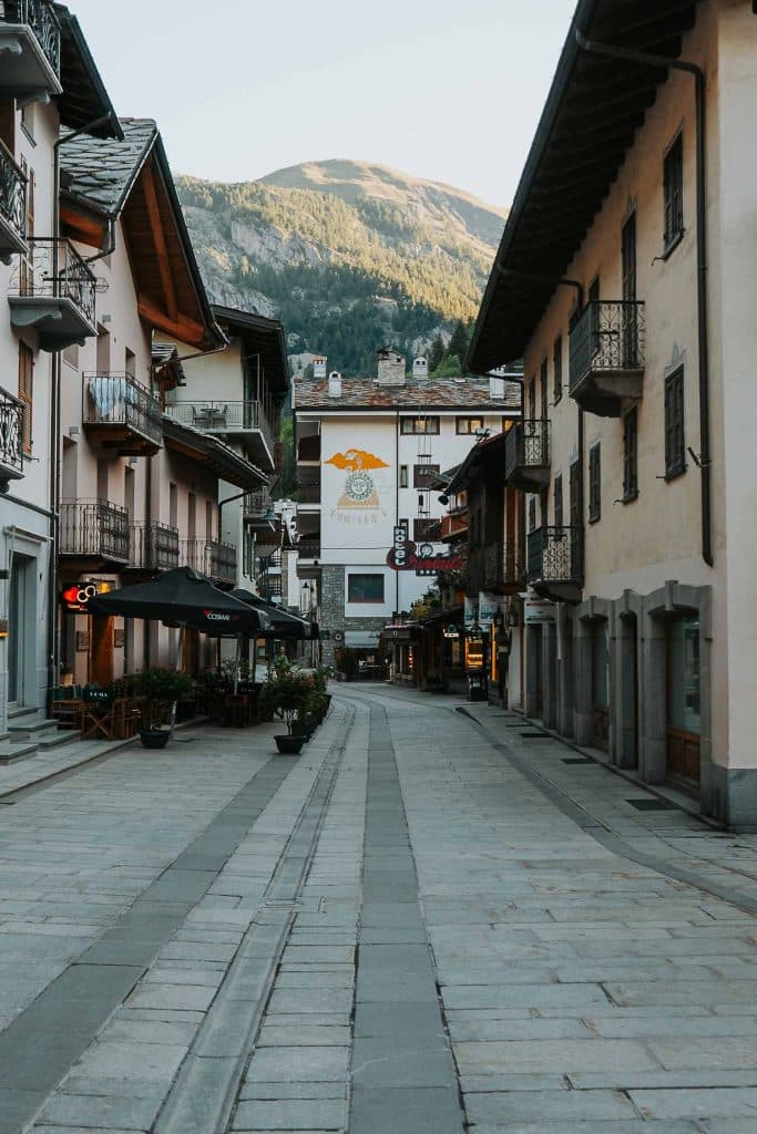 Een rustige, smalle geplaveide straat omzoomd met gebouwen en balkons in een bergdorpje, met terrasjes - perfect voor een rustige pauze tijdens een roadtrip in Valle d'Aosta - met heuvels zichtbaar op de achtergrond onder een heldere hemel.