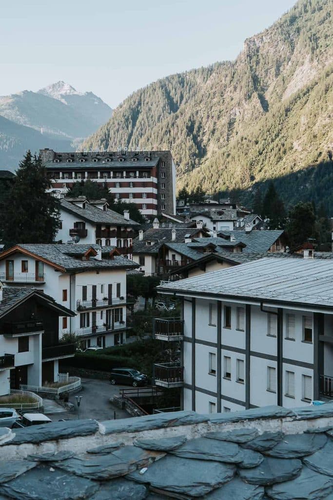 Alpendorp met witte gebouwen en grijze leistenen daken, omringd door groene beboste bergen onder een heldere hemel - een perfecte stop tijdens een roadtrip in Valle d'Aosta, met in de verte een besneeuwde top zichtbaar op de achtergrond.