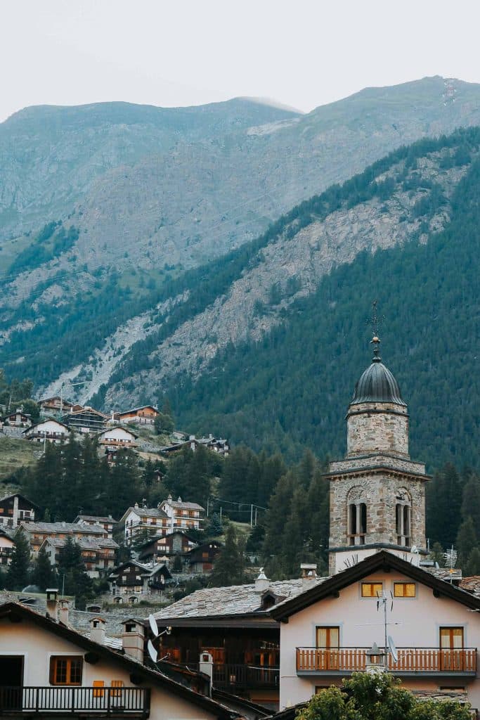 Een schilderachtig bergdorp met een stenen kerktoren op de voorgrond, traditionele huizen op de heuvel en beboste bergen op de achtergrond - een perfecte stop tijdens een roadtrip in Valle d'Aosta onder een bewolkte hemel.