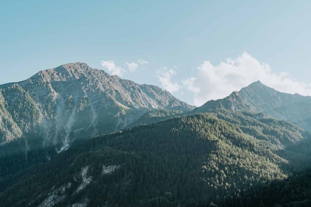 Berglandschap in Valle d'Aosta met ruige toppen en beboste hellingen onder een helderblauwe hemel met een paar pluizige wolken, alles badend in zacht daglicht-een echte Bergenmagie ontsnapping.