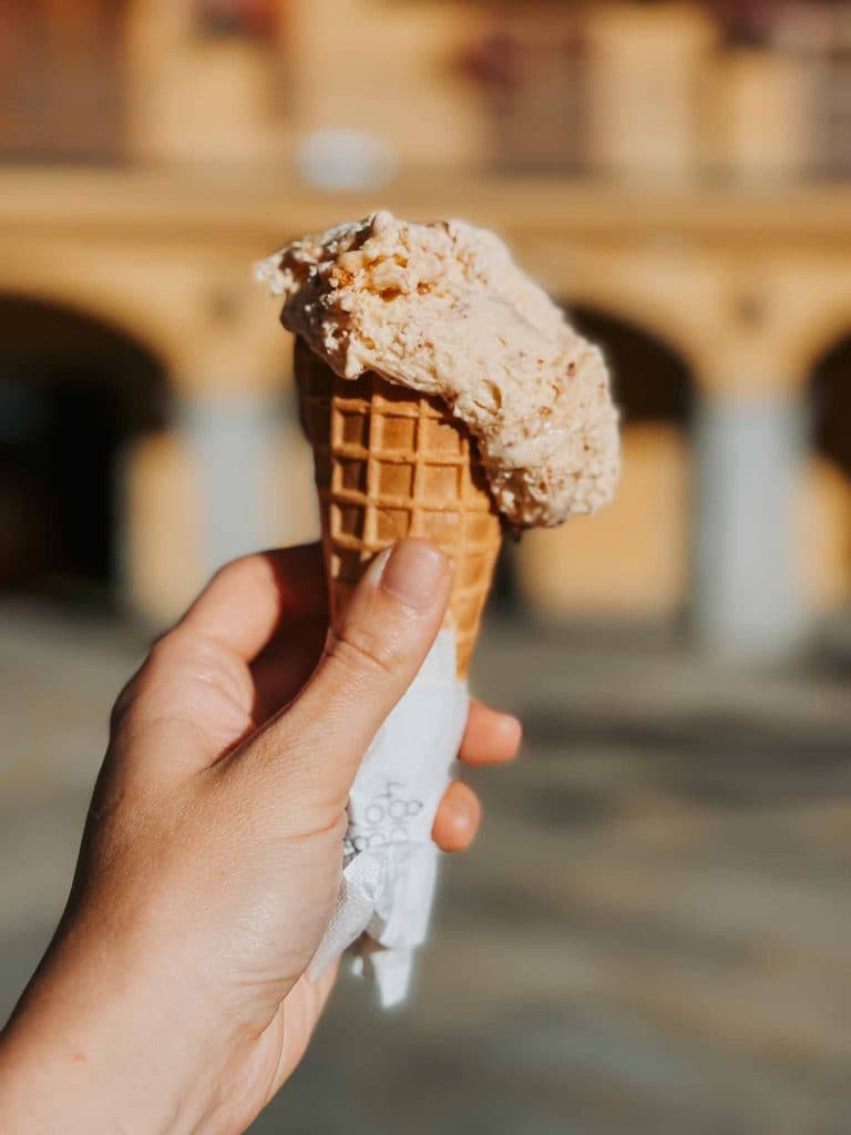 Een hand houdt een ijshoorntje vast met een bolletje lichtbruin ijs, gewikkeld in een servet, tegen een wazig gebouw - een beeld van de zoete charme van Valle d'Aosta.