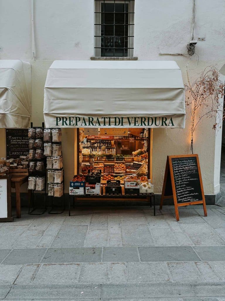 Een kleine groentewinkel met een witte luifel met de tekst "PREPARATI DI VERDURA," toont verse producten en verpakte goederen uit Valle d'Aosta. Rechts staat een krijtbordmenu en links hangen Bergenmagie-verpakkingen.