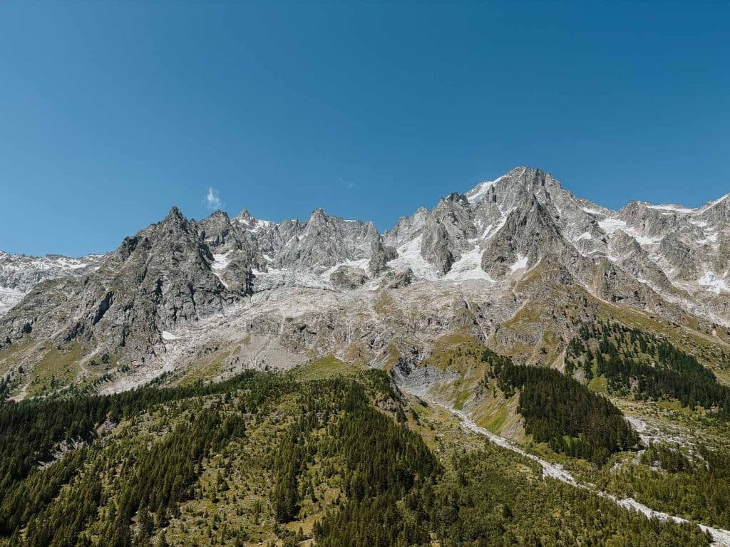 Besneeuwde rotsachtige bergtoppen rijzen op onder een helderblauwe hemel in Valle d'Aosta, met groene dennenbossen op de lagere hellingen en rotsachtig terrein zichtbaar in het midden.