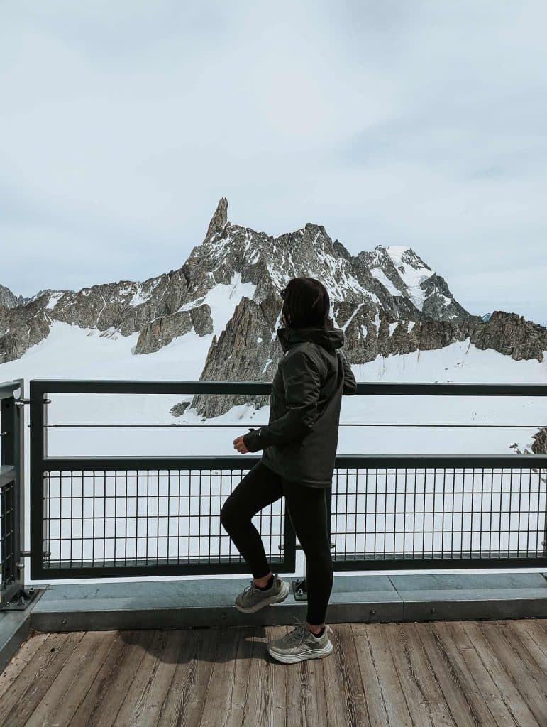 Een persoon in een donkere jas en sportschoenen staat op een houten platform en kijkt uit op het Bergenmagie van besneeuwde bergen in Valle d'Aosta, waarvan de grillige toppen oprijzen onder een bewolkte hemel.