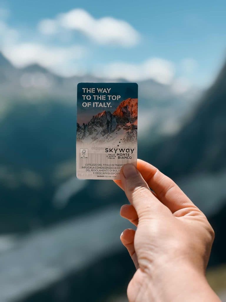 Een hand houdt een Skyway Monte Bianco ticket vast met bergen op de achtergrond. Op het kaartje staat "De weg naar de top van Italië" en er zijn besneeuwde bergtoppen te zien die de Bergenmagie en de schilderachtige alpine schoonheid van Valle d'Aosta vastleggen.