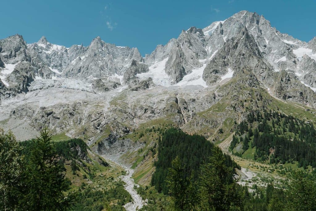 Besneeuwde, rotsachtige bergen rijzen uit boven groene bossen en een kronkelend beekje onder een helderblauwe hemel in het hart van Valle d'Aosta. Verspreid liggende sneeuw en ruig terrein creëren een dramatisch, natuurlijk landschap.
