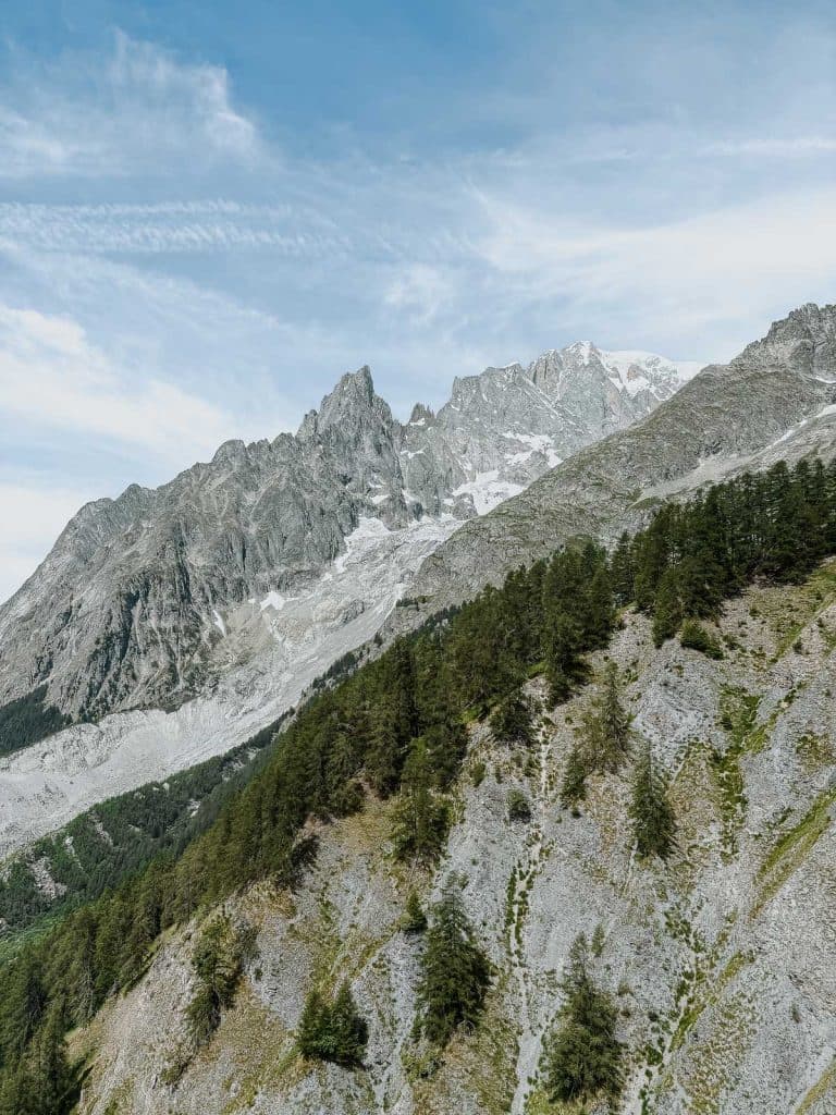 Grillige pieken van het Bergenmagiegebergte, gedeeltelijk bedekt met sneeuw, steken uit boven een beboste heuvel in Valle d'Aosta, dit alles onder een blauwe hemel met pluizige wolken.