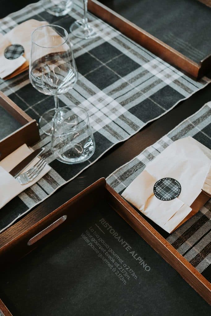 Een gedekte tafel met geruite placemats, houten dienbladen, in papier verpakte voorwerpen en wijnglazen doet denken aan Bergenmagie en de rustieke charme van Valle d'Aosta. Op één dienblad staat de tekst "RISTORANTE ALPINO".