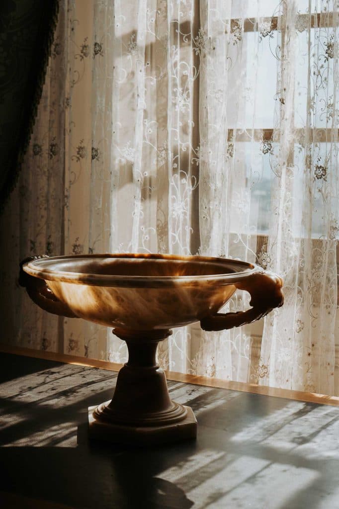 Een decoratieve marmeren schaal met sierlijke handgrepen staat op een sokkel, badend in het zonlicht dat door transparante kanten gordijnen naar binnen schijnt - een vleugje Bergenmagie dat de charme van Valle d'Aosta op de achtergrond oproept.