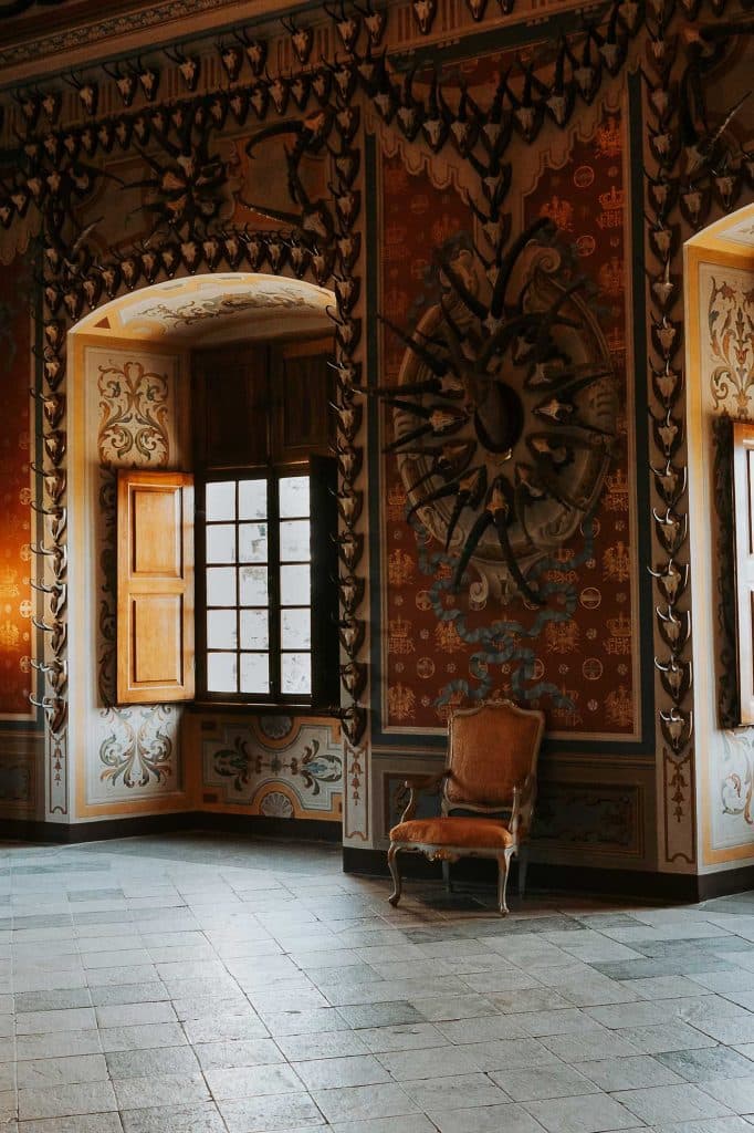 Elegante, sierlijk ingerichte kamer met hoge ramen en ingewikkelde muurontwerpen roept de charme op van Valle d'Aosta. Een oude leunstoel staat tegen de rijkelijk gedecoreerde muur onder een decoratieve zonnestraal, verlicht door zacht natuurlijk licht.