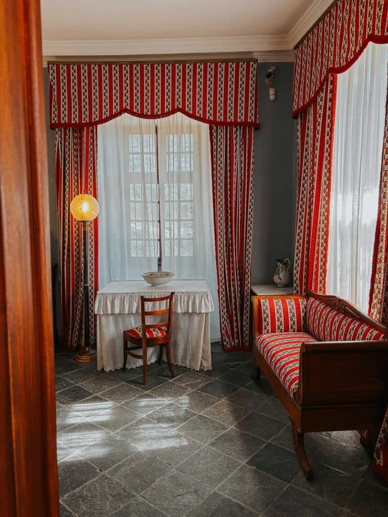 Een vintage kamer met rood en wit gedessineerde gordijnen, een bijpassende gestoffeerde bank en een vleugje Bergenmagie roept de gezellige charme van Valle d'Aosta op, terwijl het zonlicht door het raam op een met een wit kleed gedekte tafel valt.