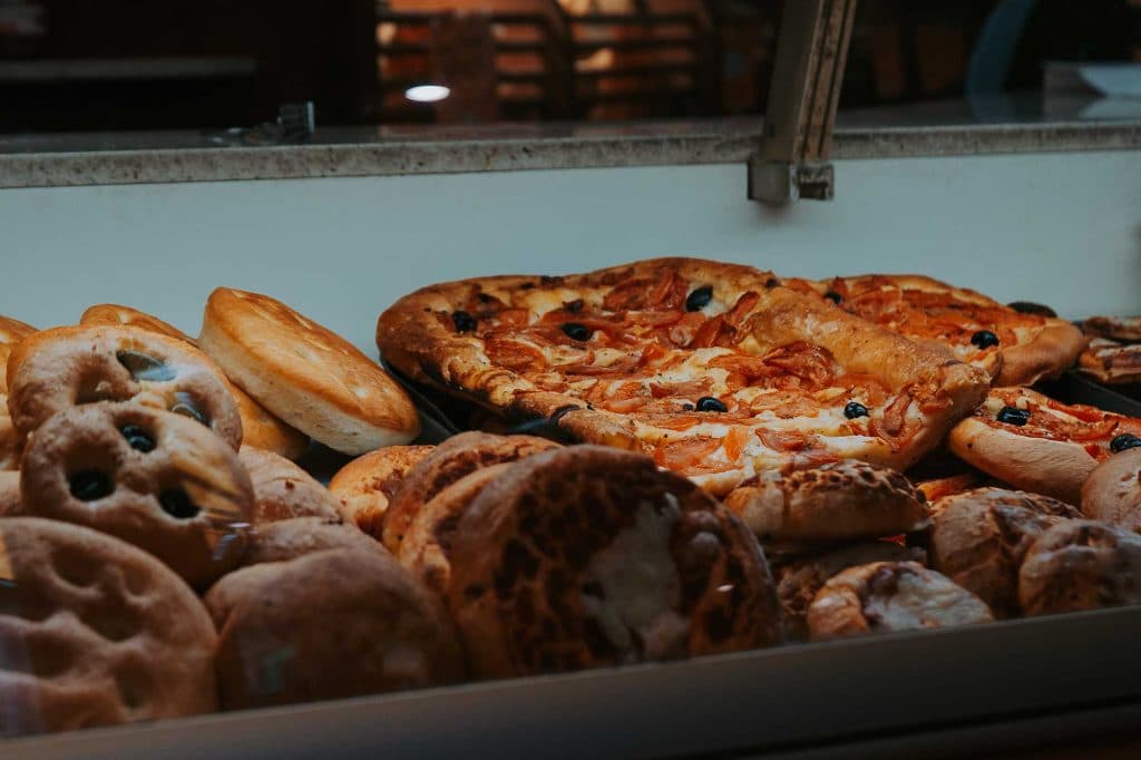 Assortiment bakkerijproducten, waaronder ronde broden en plakken pizza belegd met olijven, staan uitgestald in een glazen bakkerskast - een voorproefje van Valle d'Aosta. Op de achtergrond staan vage stoelen en tafels.