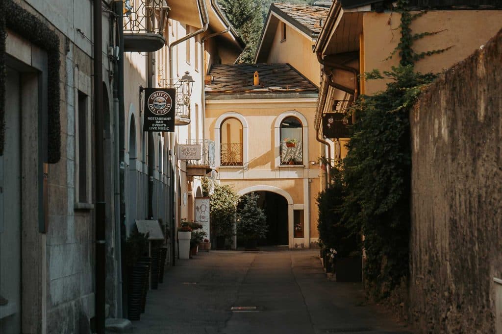 Een smal steegje met oude gebouwen leidt naar een zonovergoten geel huis met een gewelfde ingang en bloembakken. Aan de linkerkant hangt een uithangbord van restaurant Bergenmagie en tegen de rechtermuur klimt groen dat de charme van Valle d'Aosta oproept.