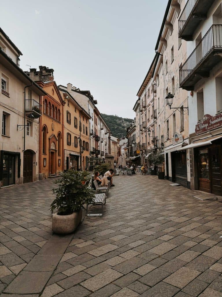 Een geplaveide straat met kleurrijke gebouwen en balkons roept de charme van Valle d'Aosta op, met mensen die ontspannen op bankjes en aan buitentafels. Heuvels en groen verheffen zich op de achtergrond onder een heldere hemel.
