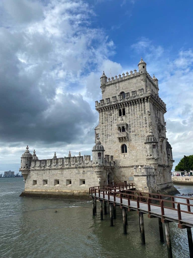 Een historische stenen toren met kantelen staat gedeeltelijk boven het water in Lissabon, verbonden met de kust door een houten loopbrug onder een blauwe wolkenhemel-een echte must-do voor bezoekers.